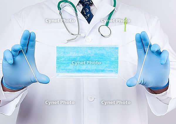 Doctor in a white coat, blue latex sterile gloves holds textile medical masks in his hand on a white background, protective accessory against viruses and bacteria [IBR125382642]