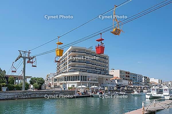 View of the Transcanal cable car with red and yellow gondolas and chairlift seats above the banks of the channeled Lez River in front of an apartment complex under a cloudless blue sky, Palavas-les-Flots, Herault Department, Occitania, France [IBR125405038]