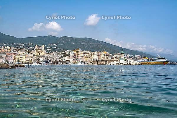 View from the clear, moving water of the small bathing area on a gravel book in the harbor near the pier of the picturesque backdrop of the Old Port of Bastia with the church of Saint-Jean-Baptiste, the green lighthouse at the pier and a ferry docked in t [IBR125405049]