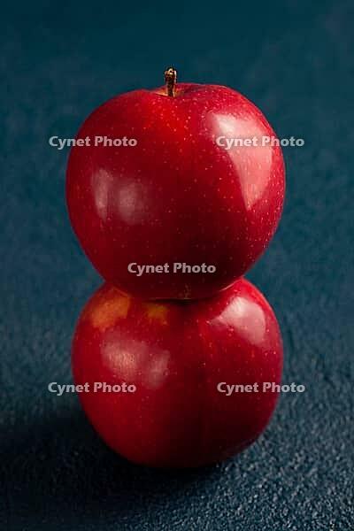 Two shiny red apples balanced stacked vertically against a textured blue background. Minimalist food concept, perfect for healthy eating, balance, or autumn-themed design and marketing visuals [IBR125405074]