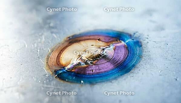 Close-up of temper color gradient bands radiating from a cooled weld point on a flat metal surface, straw to bronze to blue transition, heat affected zone, industrial macro photography, AI generated [IBR125407398]