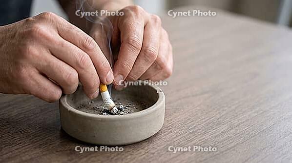 Adult hand pressing a lit cigarette into a plain unglazed ceramic ashtray on a modest laminate tabletop, thin wisp of smoke, soft natural daylight, no face visible, quitting smoking concept, AI generated [IBR125407410]