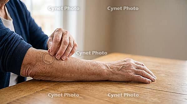 Close-up of an older adult's wrinkled hand pressing a round adhesive bandage onto the opposite forearm after vaccination, on a plain wooden table, soft natural daylight, no face visible, AI generated [IBR125407421]