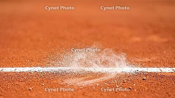 Fine white chalk dust suspended mid-air above a reddish-orange clay tennis court's boundary line, freshly disturbed mark below, bright daylight, decisive line call sports concept, AI generated [IBR125407432]