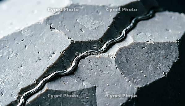 Thin metallic lithium dendrite filament tracing a jagged path along the boundary between two crystalline ceramic grains, charcoal background, industrial lighting, battery science, AI generated [IBR125407433]