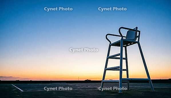 Empty white umpire chair silhouetted against a gradient dusk sky beside an unmarked tennis court, warm horizon light fading to deep blue, quiet reflective mood, sports atmosphere concept, AI generated [IBR125407440]