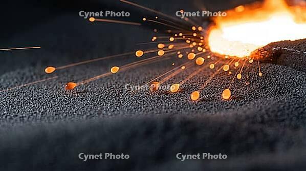 Tiny glowing orange metal spatter droplets flying from the edge of a molten weld pool above grey metal powder, frozen mid-flight, industrial macro photography, additive manufacturing, AI generated [IBR125407441]