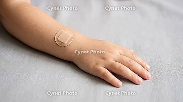 Close-up of a small child's hand and forearm with a plain round adhesive bandage after vaccination, on a soft grey fabric surface, gentle natural daylight, no face visible in frame, AI generated [IBR125407461]