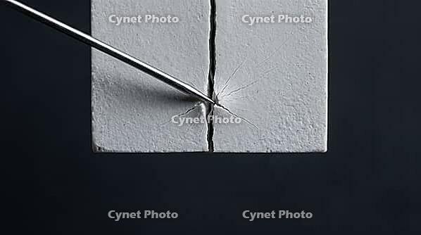 Thin metallic lithium dendrite filament tip widening a narrow crack in a pale grey ceramic block, charcoal background, wedge expansion battery failure science, AI generated [IBR125407465]