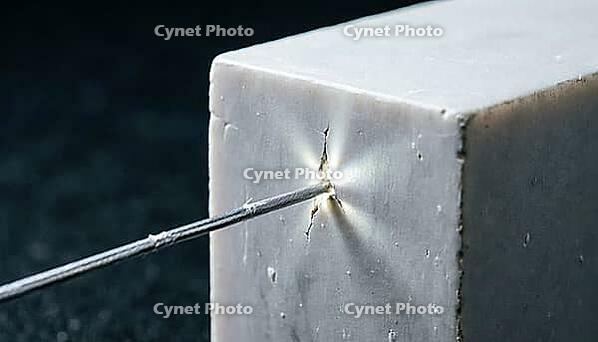Thin silver-grey metallic lithium dendrite filament piercing through a fracture point in a solid crystalline ceramic block, charcoal background, studio lighting, battery failure science, AI generated [IBR125407474]