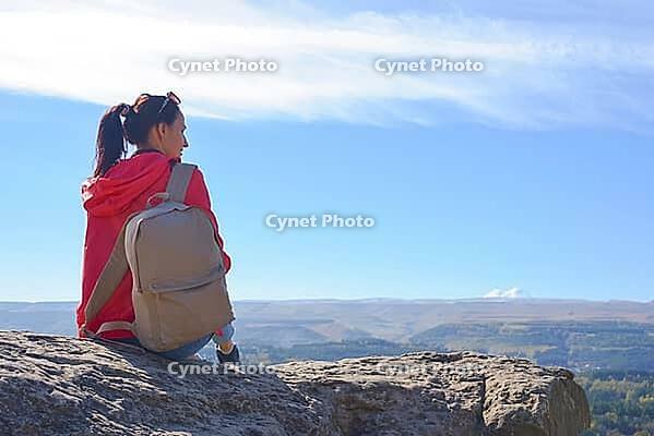 A tourist with a backpack is sitting on a mountain peak and enjoying the beautiful view. A wandering woman in an orange jacket is resting on a rock and looking out at the beautiful sunny landscape of the Caucasus [IBR125411136]