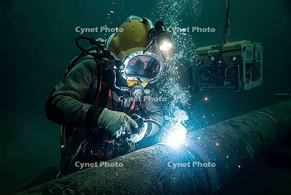Subsea hyperbaric welding on industrial gas pipeline with commercial diving helmet and rising bubbles, offshore energy infrastructure maintenance, high risk deep sea repair combining specialized human craftsmanship with robotic remotely operated vehicle s [IBR125414118]