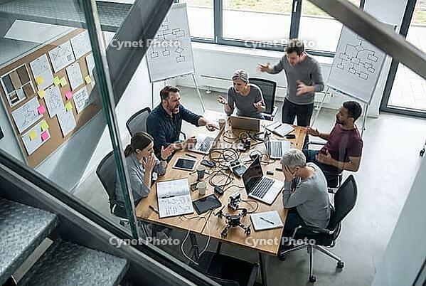 Heated workplace conflict, agile team crisis, project failure, high stress levels and burnout among engineers during chaotic hardware development, furious developers arguing intensely around a wooden conference table covered in tangled wiring, open laptop [IBR125414121]