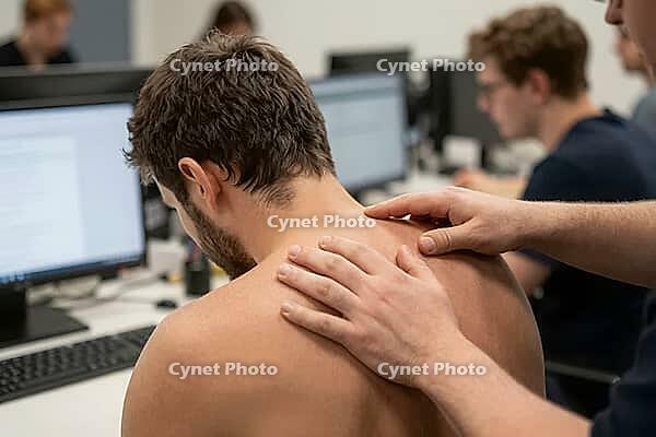 Ergonomic spine examination and occupational health physiotherapy for desk worker suffering back pain and cervical strain in digital workspace, hands of therapist gently palpating trapezius muscles and upper cervical vertebrae of shirtless young employee  [IBR125414128]