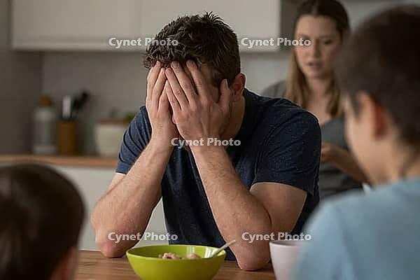 Mental breakdown and parental burnout of a desperate father sitting at breakfast table with family, emotional distress and severe anxiety overwhelm, psychological exhaustion clashes with domestic daily responsibilities, hands covering face depict chronic  [IBR125414132]