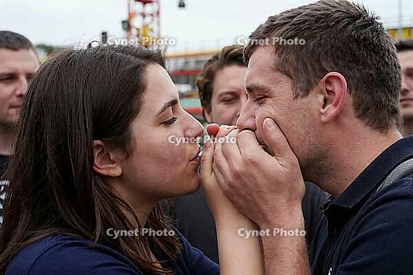 Intimate sensory connection and youth subculture during public celebration, young couple sharing breath through clasped hands in a crowd, peer group dynamics and emotional vulnerability amidst amusement park structures, closed eyes expressing profound mut [IBR125414138]