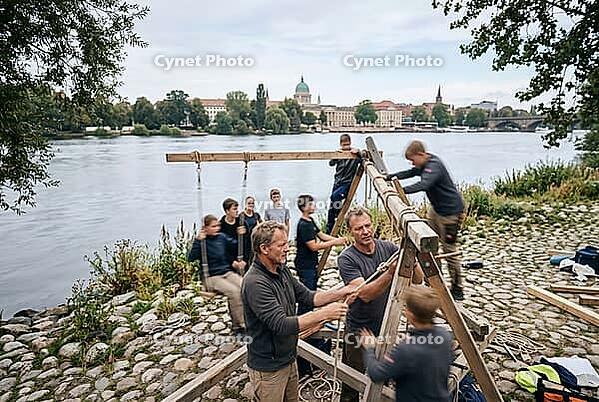 Collaborative timber construction and intergenerational craftsmanship on Potsdam riverbank, youth education outdoor workshop experiential learning, knotting ropes and stabilizing wooden beams with teamwork, hands on pedagogy fostering manual skills across [IBR125414168]