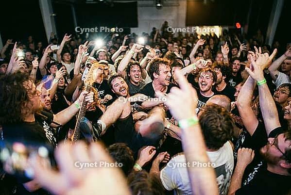 Ecstatic crowd surfing at energetic live music concert, passionate fans celebrate underground youth culture and collective euphoric togetherness, lifted audience member smiles amidst densely packed venue hall, electric bass headstock emerges from passiona [IBR125414181]
