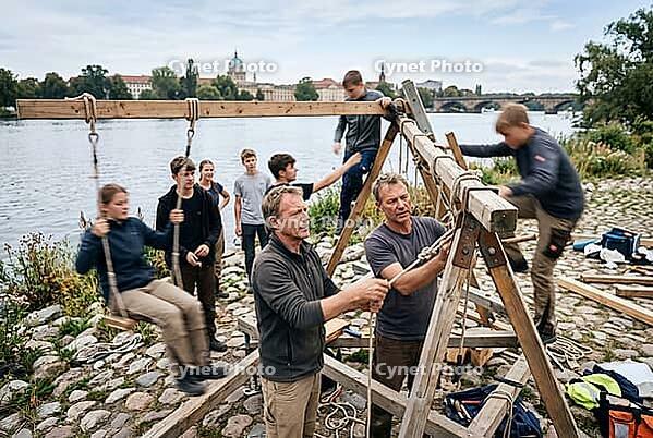 Experiential outdoor education and manual teamwork by the river, youth group and mentors constructing a wooden swing structure with ropes and timber, intergenerational knowledge transfer and vocational craft skills in social pedagogy, active collaboration [IBR125414182]