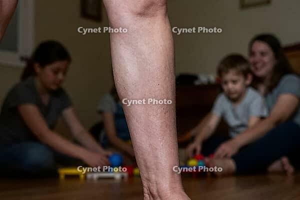 Adult leg in foreground with family and children playing in background, domestic life, generational care, parenting stability, home nursery playtime, close perspective of an adult calf and foot firmly grounded on wooden floor while children and mother pla [IBR125414214]