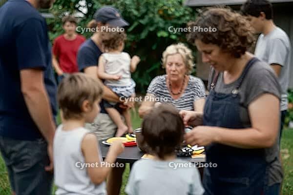 Intergenerational family cohesion and informal care economy in an outdoor domestic garden, diverse generations assemble around a shared table, grandparents alongside parents and young offspring establish social continuity, informal childcare networks mani [IBR125414216]