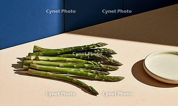 Fresh green asparagus spears rest on a beige surface. Blue wall behind adds contrast to composition. Small white plate nearby for serving or styling, AI generated [IBR125421254]