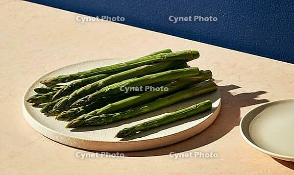 Fresh green asparagus spears on white ceramic plate. Plate rests on light beige surface. Deep blue wall behind adds contrast, AI generated [IBR125421261]