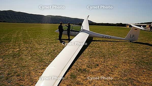 Two people standing near a glider on a grassy field with mountains in the distance on a sunny day, Aérodrome de Château-Arnoux - Saint-Auban, France [IBR125421624]