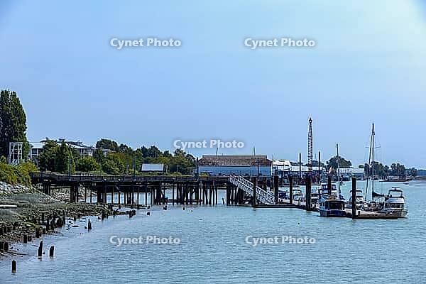 A scenic view of a waterfront with various boats moored to a wooden pier and industrial structures lining the shore, Richmond Whistler, BC, Canada [IBR125424500]