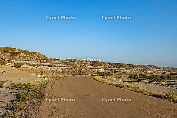 An asphalt road curves through a vast landscape of eroded hills and layered rock formations under a bright, cloudless sky, Drumheller, AB T0J 0Y0, Alberta, Canada [IBR125424505]