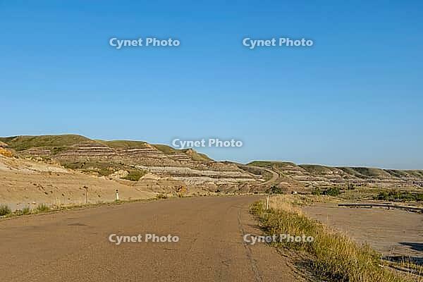 A paved road curves through a vast landscape of eroded hills displaying distinct geological strata under a bright, cloudless sky, Drumheller, AB T0J 0Y0, Alberta, Canada [IBR125424515]