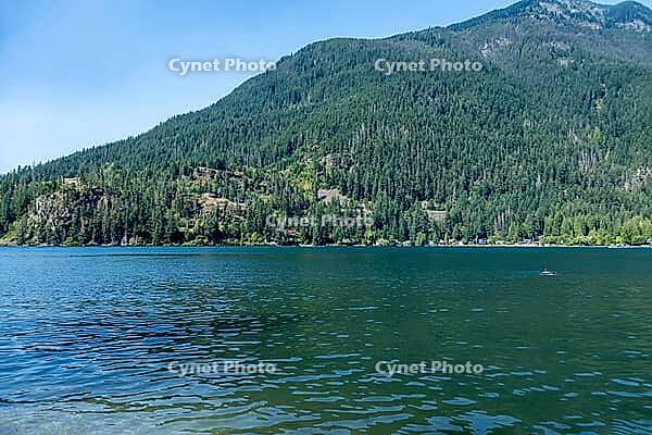A serene natural landscape featuring clear blue lake water leading to a densely forested mountain under a bright sky with scattered houses along the shore, kettle Valley trail, BC Canada [IBR125424525]