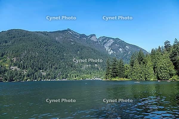 A vast, calm lake reflects the surrounding dense evergreen forests and majestic mountains under a bright, cloudless sky, kettle Valley trail, BC Canada [IBR125424539]