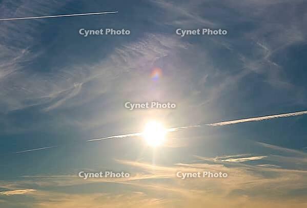 Sun illuminates cirrus clouds and airplane contrails against a blue sky, creating a lens flare [IBR125424553]