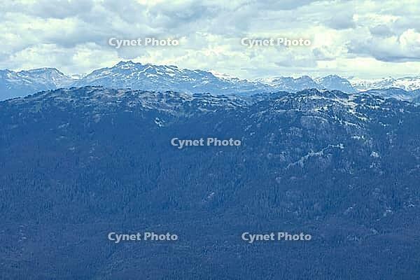 Expansive view of rugged mountain peaks with patches of snow, receding into a hazy horizon above a deep blue coniferous forest, Glacier Whistler, BC, Canada [IBR125424558]