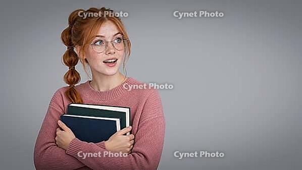 A redheaded student with freckles and round glasses holds two books against a pink sweater, looking upward with an animated expression against a gray studio setting. Cute smile, AI generated [IBR125424579]
