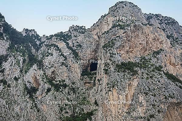 Rocky mountain cliff face displaying a prominent cave opening against a clear sky [IBR125436240]