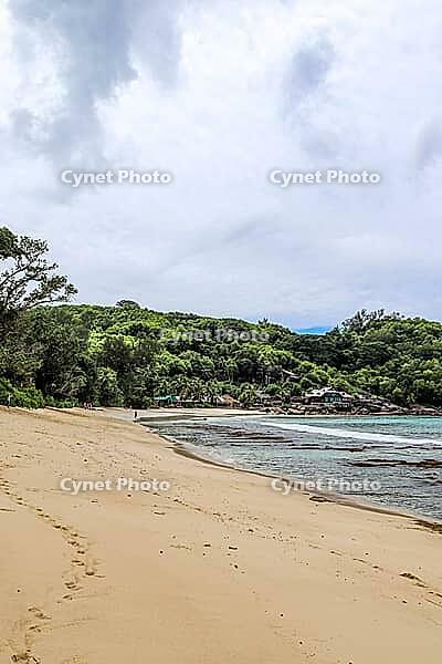 Sandy beach on tropical island showing turquoise ocean water, green jungle, and hillside architecture under cloudy sky [IBR125436258]