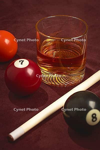 A glass of whiskey sits next to three billiard balls numbered 5, 7, and 8 on a dark table. A white cue stick lies across the table. This scene captures a moment of leisure [IBR125437990]