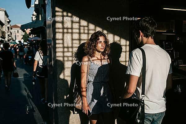 Geometric grid shadows fall upon a contemplative young woman during an emotional conversation with a man on a bustling European city street in summer, architectural latticework projections cast a psychological cage across her face evoking themes of emotio [IBR125446413]