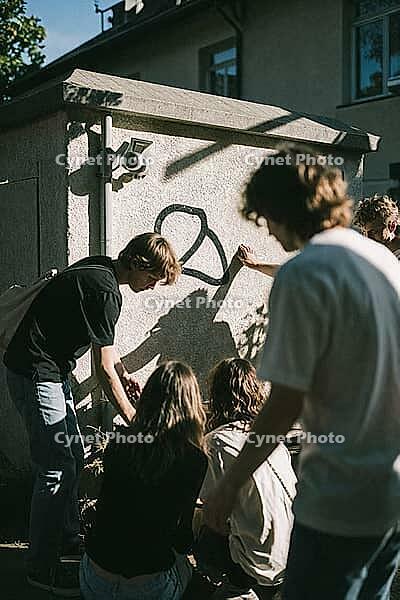 Youth subculture and communal connection thrive outdoors, young adults gathering near urban walls with surveillance cameras and graffiti artwork, candid camaraderie merges with societal tension beside a utility pavilion, harsh diagonal afternoon sunlight  [IBR125446419]
