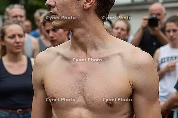 Athletic shirtless young man stands amidst a dense public crowd outdoors during summer, displaying physical presence, vulnerability, exposed muscular torso, attentive onlookers observing the gathering, collective gaze focusing on individual strength, anat [IBR125446430]