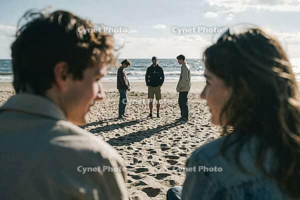 Interpersonal connection among young adults on sandy coastal terrain under harsh daylight reveals diverse group dynamics and thoughtful reflections, two blurry figures in the foreground frame three distant companions standing in deep conversation near bre [IBR125446437]