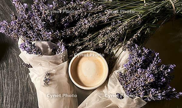 Cup of coffee sits surrounded by dried lavender sprigs. Lavender flowers rest on a dark wooden surface. White tissue paper wraps some lavender bundles, AI generated [IBR125446439]