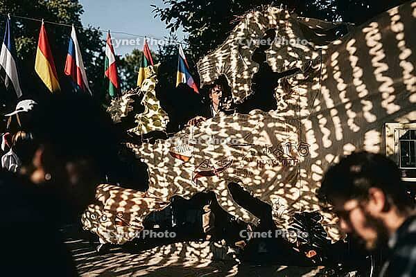 Performer behind a large cutout textile map of Europe during an outdoor theatrical event, dynamic dappled sunlight creating intricate shadow patterns across continental landmasses, vibrant international national flags suspended along a tension wire in the [IBR125446443]