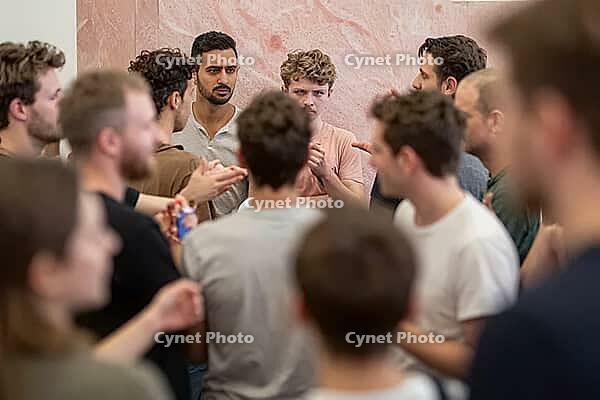 Dynamic group debate and social interaction among diverse young men indoors, expressive faces convey intense concentration, skepticism and dialogue, diverse individuals assemble in close proximity against a polished pink stone wall, shallow depth of field [IBR125446449]
