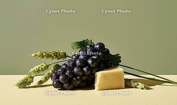 Dark purple grapes sit beside a wedge of hard cheese. Green wheat stalks rest nearby. Soft light highlights textures, AI generated [IBR125446450]