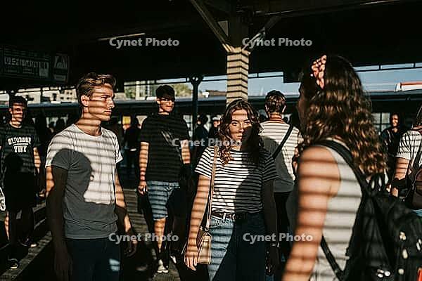 Commuters traverse railway platform during rush hour, rhythmic slatted shadows cast across young travelers standing in contemplation, transit passengers experiencing quiet anticipation within public transport networks, warm sunlight filtering through stat [IBR125446454]