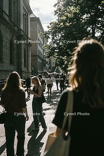Pedestrians navigate busy urban sidewalks bathed in intense summer sunlight and contrasting shadow, mobile phone distraction reflects modern digital isolation, historical stone architecture flanks the crowded thoroughfare while leafy canopy foliage filter [IBR125446456]