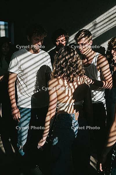 Subtle emotional tension, vulnerability and group cohesion unfold among young adults gathered in dramatic natural window light through blinds, casting parallel shadow stripes across bodies and bare skin, introspective glances and contemplative facial expr [IBR125446464]